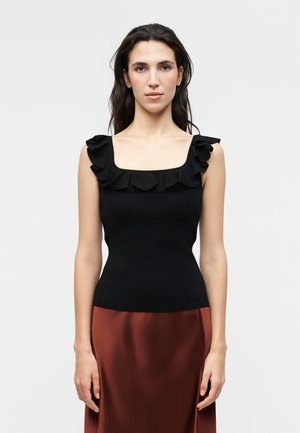 Γυναίκα με μακριά σκοτεινά μαλλιά, που φοράει αμάνικο μαύρο τοπ με βολάν στο τετράγωνο ντεκολτέ και καφέ σατέν φούστα, στέκεται μπροστά σε απλό φόντο.