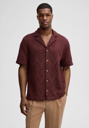 Mann trägt ein kurzärmeliges, kastanienfarbenes Hemd mit Knöpfen und Zickzackmuster sowie eine beige Hose, steht vor einem schlichten hellgrauen Hintergrund.