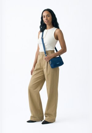 Hosszú sötét hajú nő fehér ujjatlan felsőben, bézs színű, bőszárú nadrágban, fekete cipőben, és egy kis kék válltáskával a vállán.