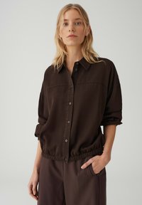 Donna con capelli biondi che indossa una camicia marrone scuro abbottonata e pantaloni coordinati, una mano in tasca, espressione neutra, sfondo chiaro e uniforme.