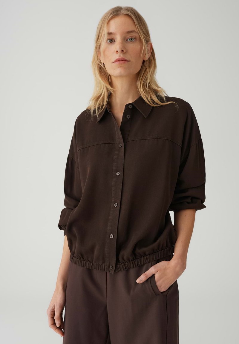 Donna con capelli biondi che indossa una camicia marrone scuro abbottonata e pantaloni coordinati, una mano in tasca, espressione neutra, sfondo chiaro e uniforme.