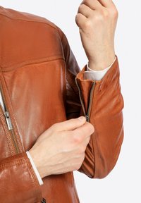 Giacca di pelle marrone con polsini con zip, texture liscia, vestibilità su misura e dettagli di cucitura sottili.