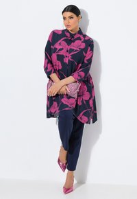 Blumige Tunika in Marineblau mit pinken Blumenmustern, kombiniert mit Marineblauen Hosen und glänzenden pinken High Heels. Acryltasche in der linken Hand gehalten.