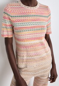 Haut en tricot présentant des motifs zigzag multicolores en rose, orange, vert et crème, avec des manches courtes et un col rond.
