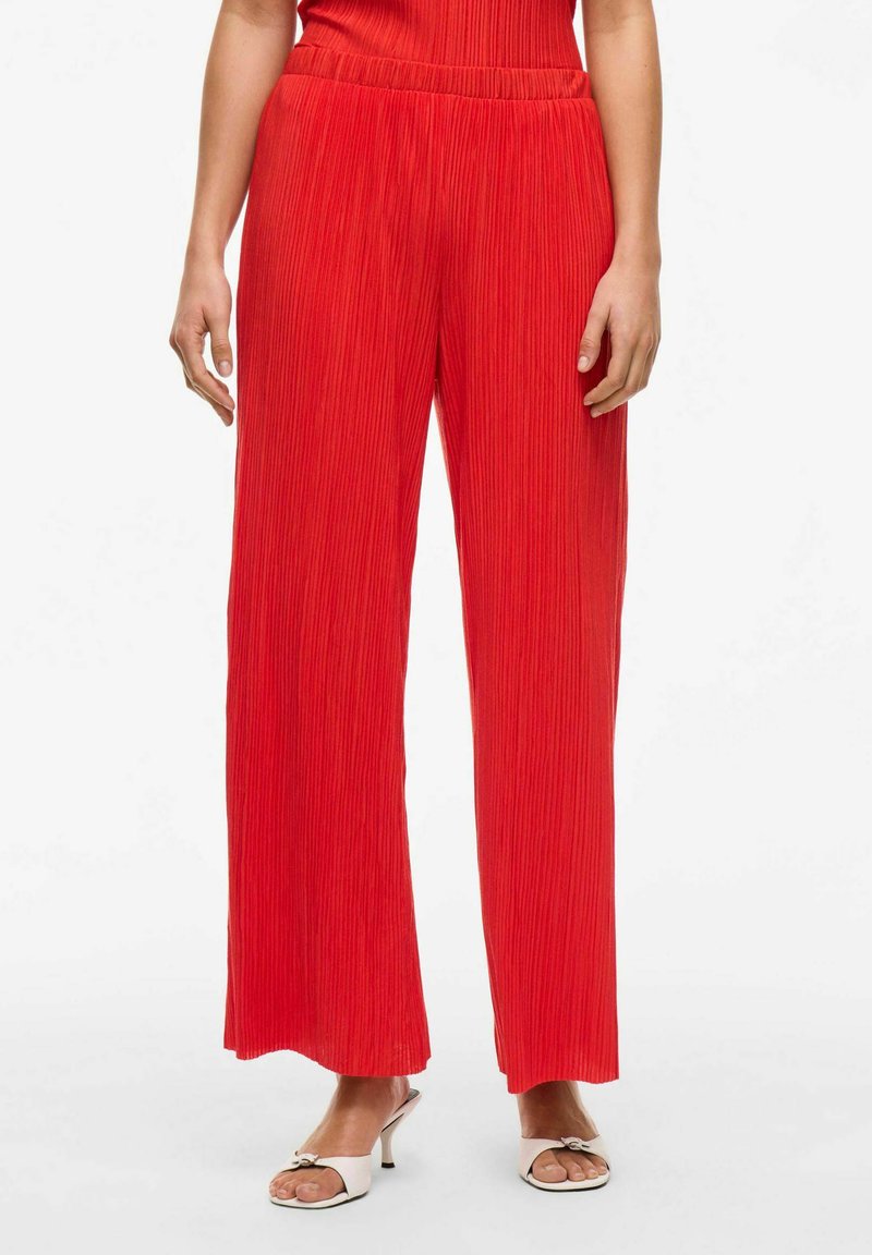 Pantalones anchos de pierna amplia en rojo, con pliegues y una cintura elástica. Textura suave y tela ligera. Se llevan con zapatos blancos de punta abierta.