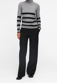 Pull à col roulé gris avec des rayures noires, des poignets côtelés et une coupe ajustée, associé à un pantalon noir à jambes larges et des chaussures noires.