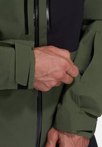Veste verte et noire avec un fini texturé, dotée d'une poche zippée sur la manche et de poignets réglables avec un système de fermeture sécurisé.