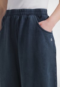 Osoba ubrana w granatowe spodnie z elastycznym pasem, z jedną ręką w bocznej kieszeni, na tle jednolitego jasnego tła.