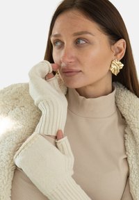 Rękawiczki bez palców z kremowego prążkowanego splotu, noszone z beżową bluzką z półgolfem oraz dużymi teksturowanymi złotymi kolczykami, na tle jednolitej białej ściany.