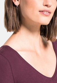 Top de pico en tejido color burdeos profundo, que muestra una textura suave. Modelo con pequeños pendientes de aro de plata, con hombros y cuello visibles.