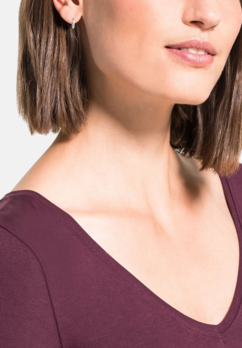 Top de pico en tejido color burdeos profundo, que muestra una textura suave. Modelo con pequeños pendientes de aro de plata, con hombros y cuello visibles.