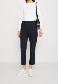 Bílý top s dlouhým rukávem, tmavě modré kalhoty s elastickým pasem, bílé nazouvací boty a malá černá kabelka přes rameno. Jednoduchý design a pohodlný střih.