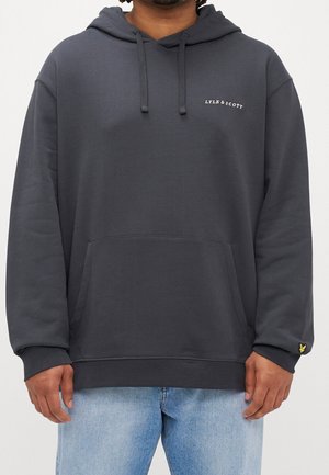 Mann trägt dunkelgrauen Lyle & Scott Hoodie mit Fronttasche, Kordelzug und gelbem Logo am Ärmelbündchen, kombiniert mit hellblauen Jeans.