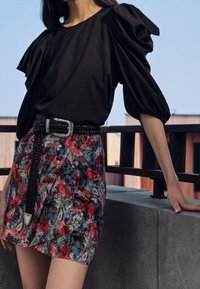 Maglietta nera a maniche a sbuffo abbinata a una mini gonna a fiori con tonalità rosse e blu, accentuata da una cintura nera intrecciata.