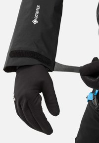 Gant en Gore-Tex noir avec un ajustement serré, présentant une texture lisse. Une bande Velcro attachée et une boucle de tension sont visibles au niveau du poignet.