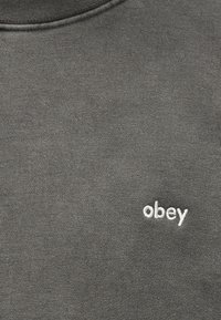 Σκούρο γκρι ύφασμα με μικρό λευκό κεντημένο κείμενο «obey» στην πάνω αριστερή πλευρά, πιθανώς φούτερ ή μπλουζάκι.