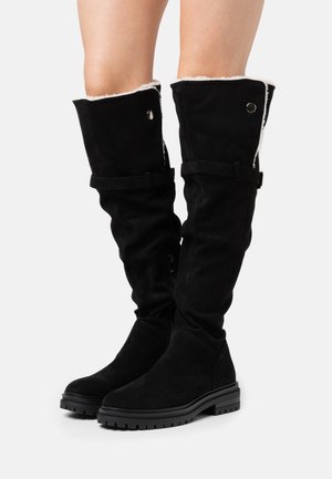 Botas negras por encima de la rodilla hechas de ante con un forro suave. Incluyen una suela de goma baja, correas ajustables y detalles en plata.