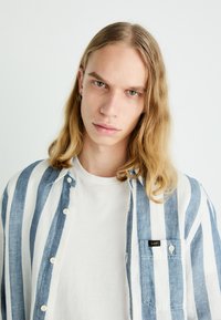 Gestreifte Button-Down-Bluse in Blau und Weiß, aus leichtem Stoff gefertigt, über einem einfachen weißen T-Shirt getragen, mit einer Brusttasche und Logo.