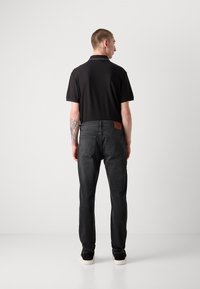 Schwarzes Polo-Shirt und dunkelgraue Jeans mit einem Leder-Patch auf der Rückseite, Slim-Fit-Design und einer sauberen, glatten Textur. Schwarze Sneakers runden das Outfit ab.