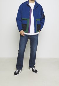 Blå zip-jacka med gröna och lila accessoarer, två framfickor och nätdetaljer. Bärs över en vit t-shirt och blå jeans, med svarta skor.