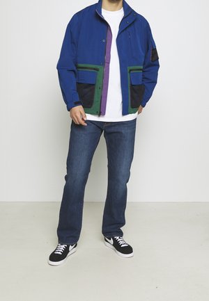 Giacca blu con zip, dettagli verdi e viola, due tasche frontali e inserti in rete. Indossata sopra una t-shirt bianca e jeans blu, con scarpe nere.