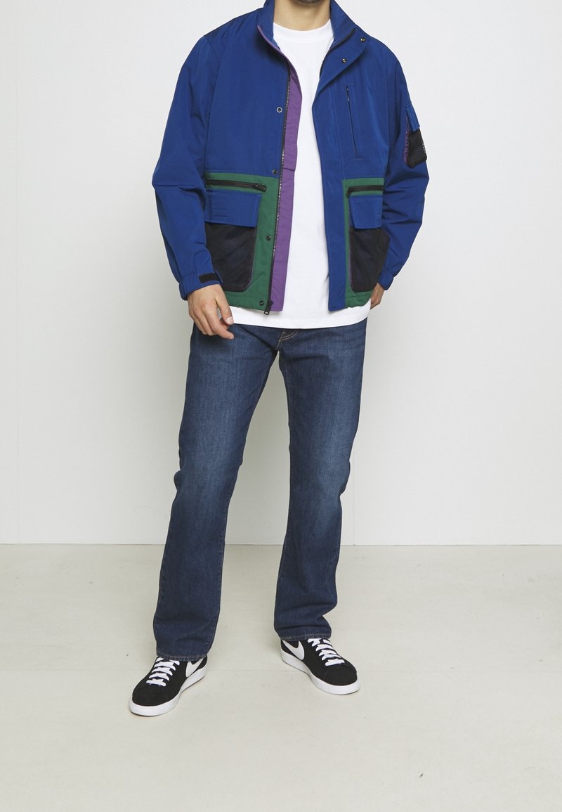 Blå zip-jacka med gröna och lila accessoarer, två framfickor och nätdetaljer. Bärs över en vit t-shirt och blå jeans, med svarta skor.