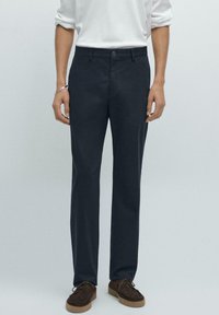 Tmavě námořnické kalhoty vyrobené z bavlněné směsi, s úzkým střihem, rovním předním dílem a jemným švem. Nosené s bílou košilí s dlouhým rukávem a hnědými botami.