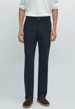 Mörkblå byxor i en bomullsblandning, med smal passform, platt framsida och subtila sömmar. Bärs med en vit skjorta med långa ärmar och bruna skor.