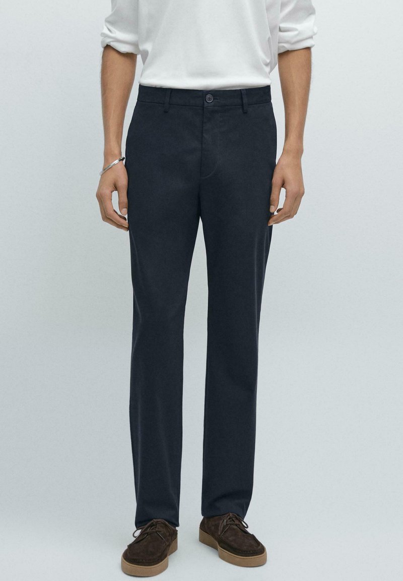 Tmavě námořnické kalhoty vyrobené z bavlněné směsi, s úzkým střihem, rovním předním dílem a jemným švem. Nosené s bílou košilí s dlouhým rukávem a hnědými botami.