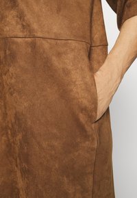 Main reposant dans la poche d'un vêtement en daim marron avec des coutures visibles et un tissu texturé.