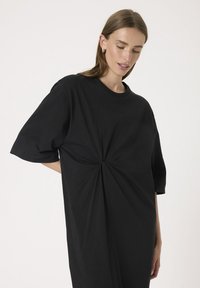 Svart oversized t-shirt klänning gjord av mjuk bomull, med korta ärmar och ett vridet designelement framtill för extra detalj.