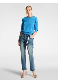 Maglione azzurro chiaro glitterato con maniche a 3/4, abbinato a jeans blu sbiaditi. Sandali a tacco alto dorati e una sciarpa fantasia nella tasca dei jeans.