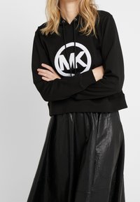 Felpa nera corta con un logo circolare bianco ‘MK’ sul davanti, abbinata a una gonna nera brillante e plissettata. Ci sono dei lacci sul cappuccio.