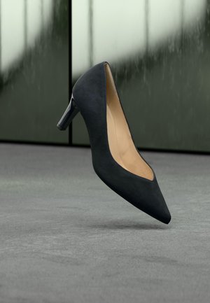 Sort spids tå højhælet sko hængende over en grå gulv med en sløret grønlig-grå baggrund.