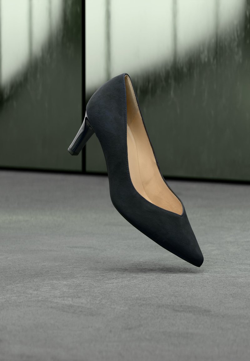 Μαύρο ψηλοτάκουνο παπούτσι με μυτερή μύτη, αιωρούμενο πάνω από γκρι δάπεδο με θολό πράσινο-γκρι φόντο.