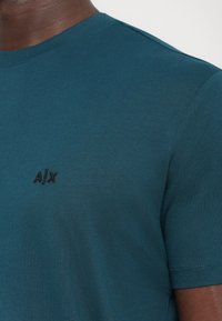 Türkiz színű, rövidujjú póló, puha anyagból készült, kerek nyakkivágással és egy kis fekete hímzett „A/X” logóval a mellkasán.