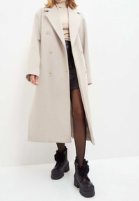 Abrigo beige de doble botonadura con solapas, mangas largas y dos botones. Lleva puesto sobre una blusa ajustada y una falda corta negra con medias transparentes.