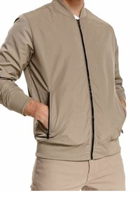 Homme portant une veste bomber beige avec une fermeture éclair noire et des poches latérales zippées, les mains dans les poches, assortie à un pantalon beige.