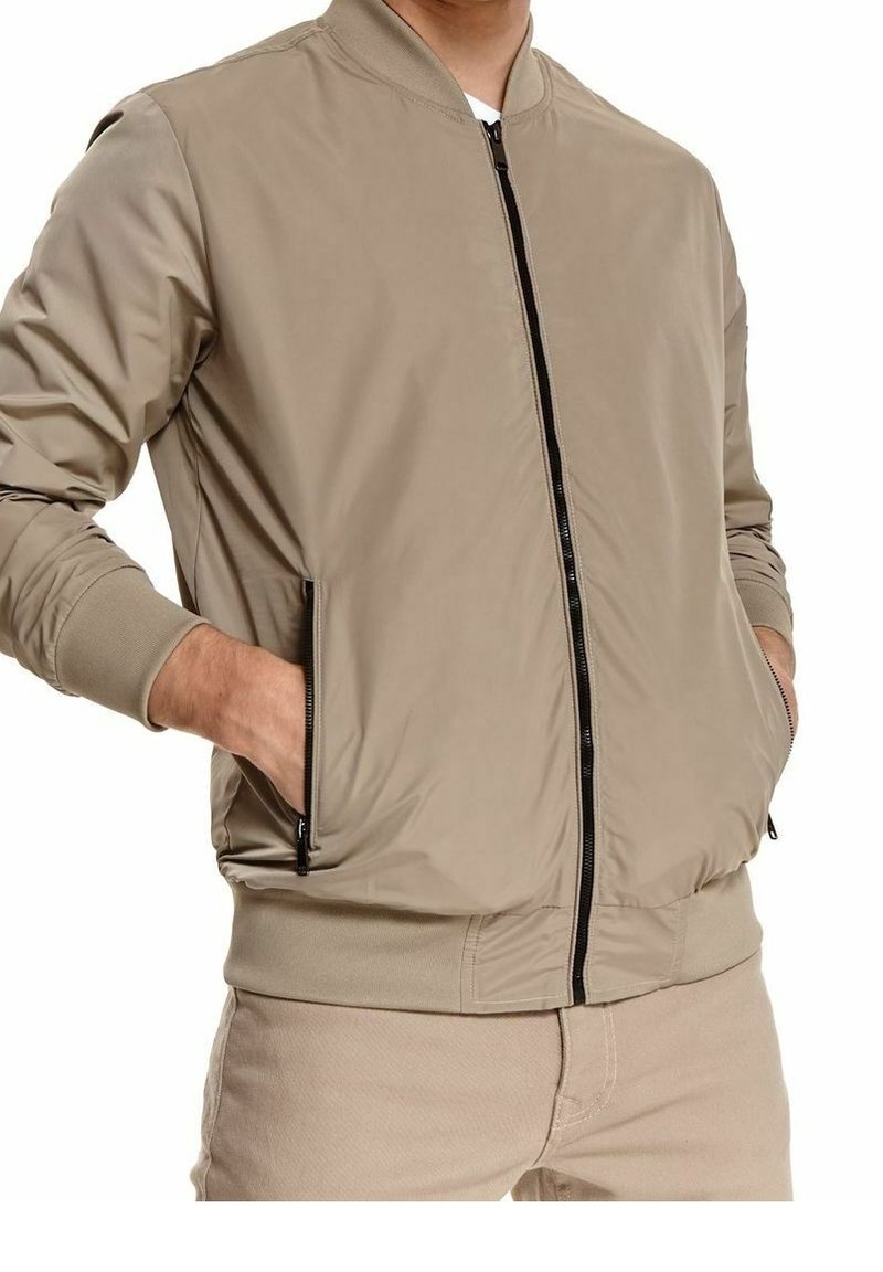 Homme portant une veste bomber beige avec une fermeture éclair noire et des poches latérales zippées, les mains dans les poches, assortie à un pantalon beige.