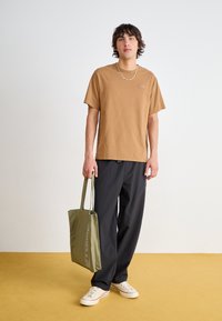 Braunes Baumwoll-T-Shirt mit Logo, schwarze lässige Hose, weiße Leinen-Sneaker und eine grüne Tragetasche mit Text. Gelber Hintergrund.