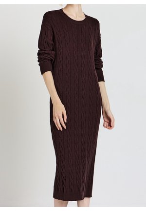 Personne portant une longue robe-pull à torsades marron foncé, manches longues retroussées, debout devant un fond clair uni.