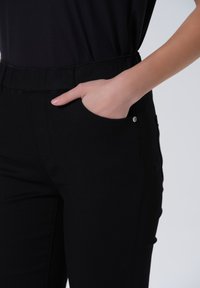 Zwarte broek met een elastische tailleband, voorzien van een zijzak en een kleine metalen accent. Het materiaal lijkt glad en rekbaar.