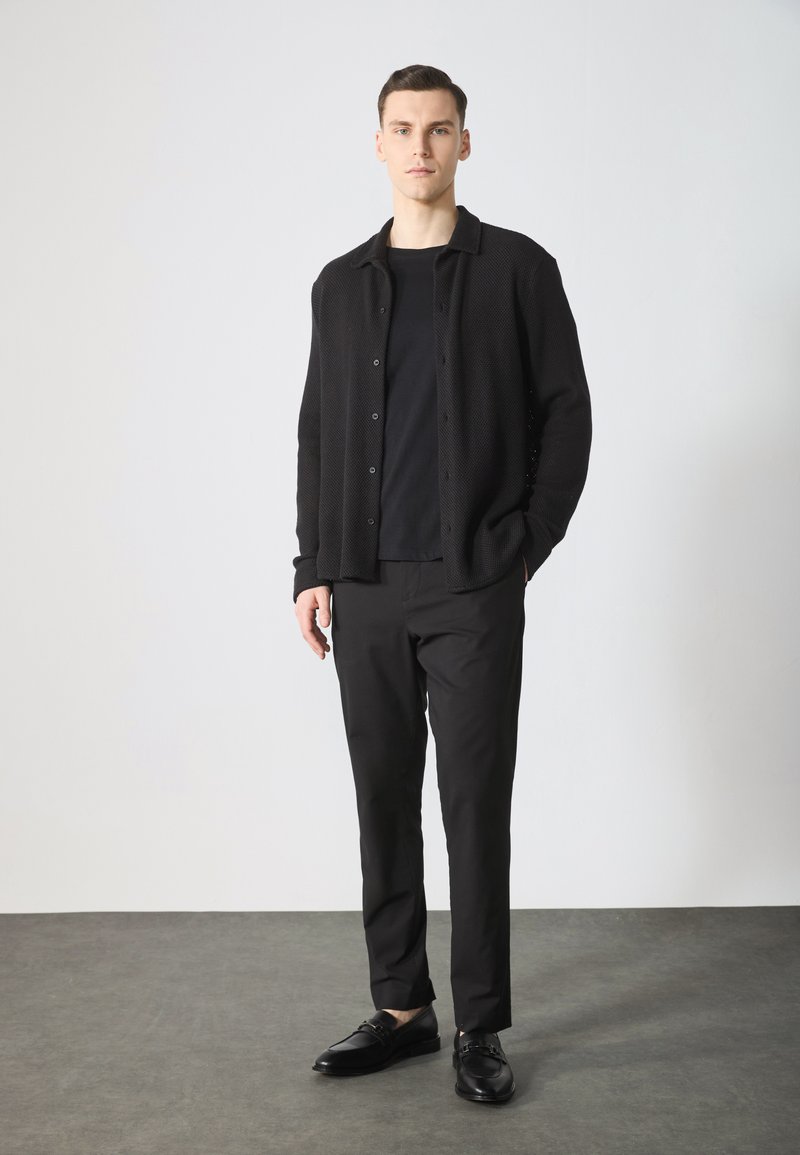 Cardigan nero lavorato a maglia con bottoni, indossato sopra una camicia nera, abbinato a pantaloni neri e scarpe nere, il tutto su uno sfondo neutro.