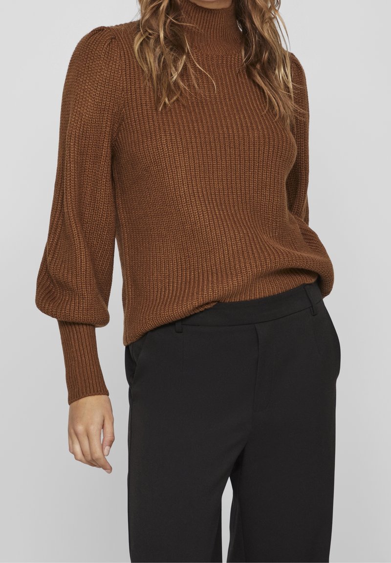 Femme portant un pull en maille côtelée marron avec des manches bouffantes et un pantalon noir taille haute sur un fond uni.