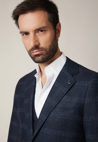 Granatowa marynarka w kratę z mieszanki wełny z subtelnym wzorem. Posiada klapę w szpic, pojedynczą kieszeń na piersi oraz srebrny detal z logo.