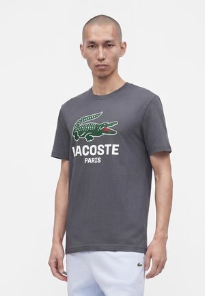 Homme portant un t-shirt gris Lacoste avec un grand logo crocodile vert et un texte blanc, associé à un pantalon blanc.