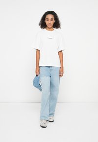 White oversized t-shirt with black logo, paired with light blue denim jeans and a blue cap. Shoes feature grey and black design.