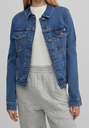 Person iført blå denimjakke over hvid strikket sweater og lysegrå joggingbukser med elastik i taljen.
