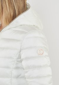 Giacca a vento bianca con cappuccio, caratterizzata da un design trapuntato, una superficie lucida e un patch circolare sul braccio sinistro con un logo.