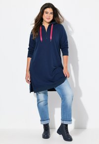Abito a felpa blu/navy con collo a zip, lacci rosa, maniche lunghe e spacchi laterali, abbinato a jeans azzurri arrotolati e stivali neri.